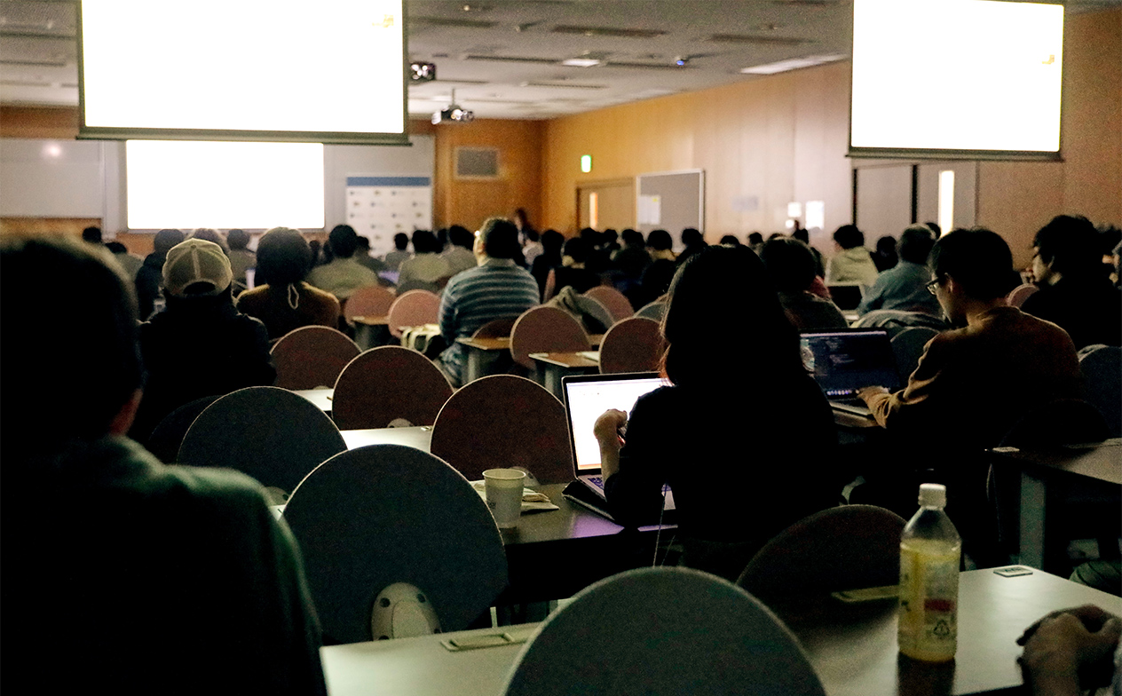 写真：スライドが前方1つ・後方2つあるルームAにて照明を暗いなか、セッション・スライドに集中している参加者の様子