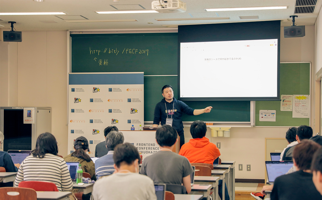 写真：ワークショップの様子。黒板を背に講師の井関さんが教卓に立って説明している。参加者は教室の各席にてノートPCを開き説明を受けている。